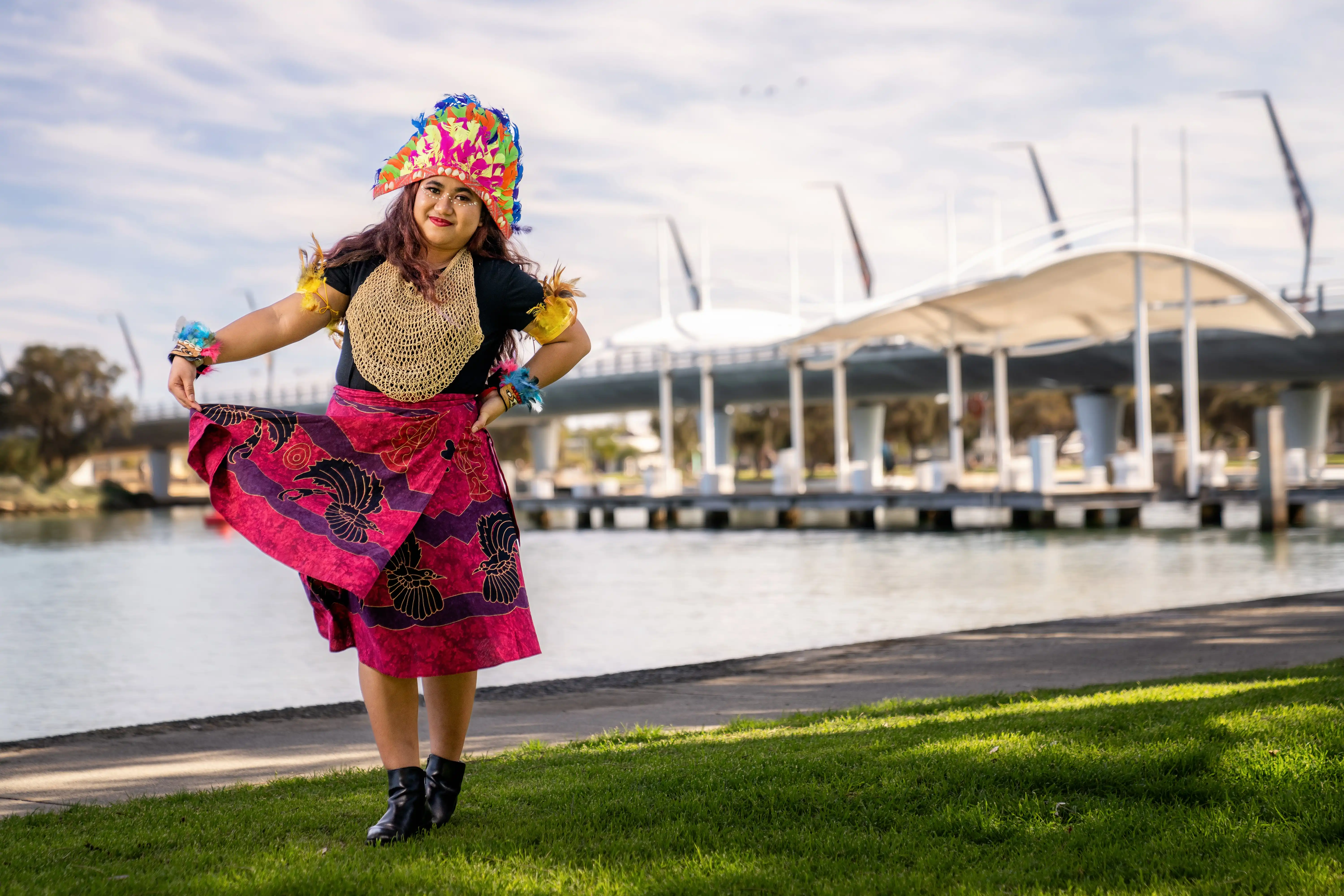 Person wearing a colourful headdress and vibrant patterned outfit poses outdoors near a waterfront